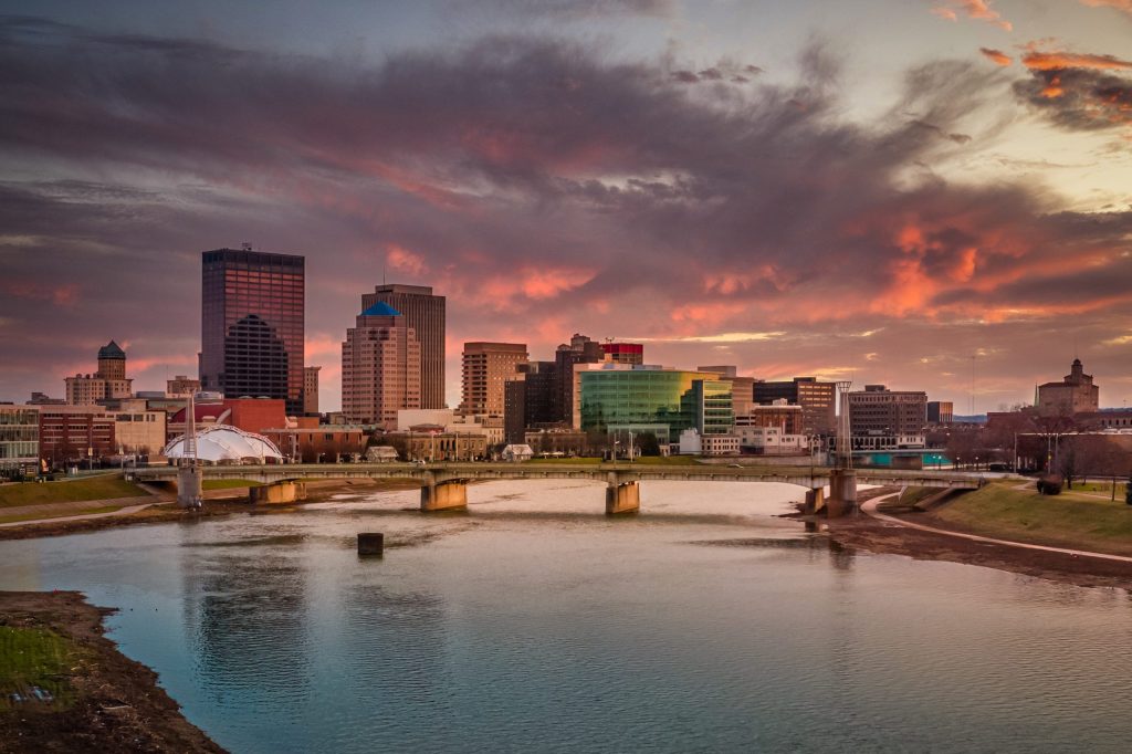 The skyline of downtown Dayton, Ohio under a sunset of pink clouds from the Great Miami River taken from a drone for an aerial view.